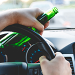 close-up of a person’s hands inside a car. One hand is gripping the steering wheel, while the other holds a green bottle, likely containing alcohol